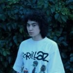 Person in white graphic tee standing outdoors with leafy backdrop.