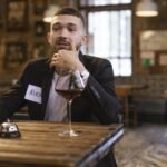 A man with a name tag sits at a table in a rustic bar environment holding a drink.